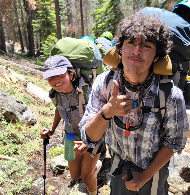 Two young hikers are captured in a sunny, wooded area. The male hiker on the right gives a thumbs-up, wearing a plaid shirt and backpack. The female hiker on the left, sporting a cap and backpack, holds a hiking stick and a water bottle. Both appear to be enjoying their outdoor adventure.